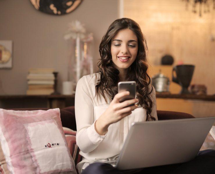 woman sitting on sofa while looking at phone with laptop on lap