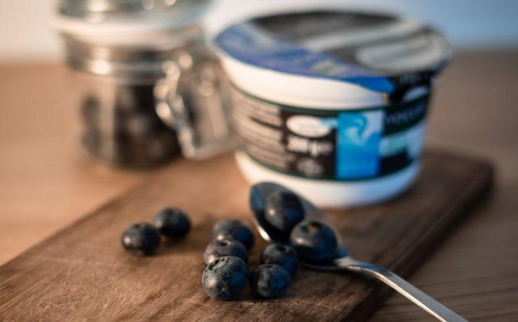 a wooden cutting board topped with blueberries next to a jar of yogurt