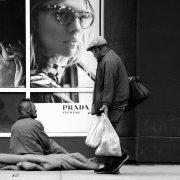 grayscale photo of man with shop bags walking past beggar siting on sidewalk